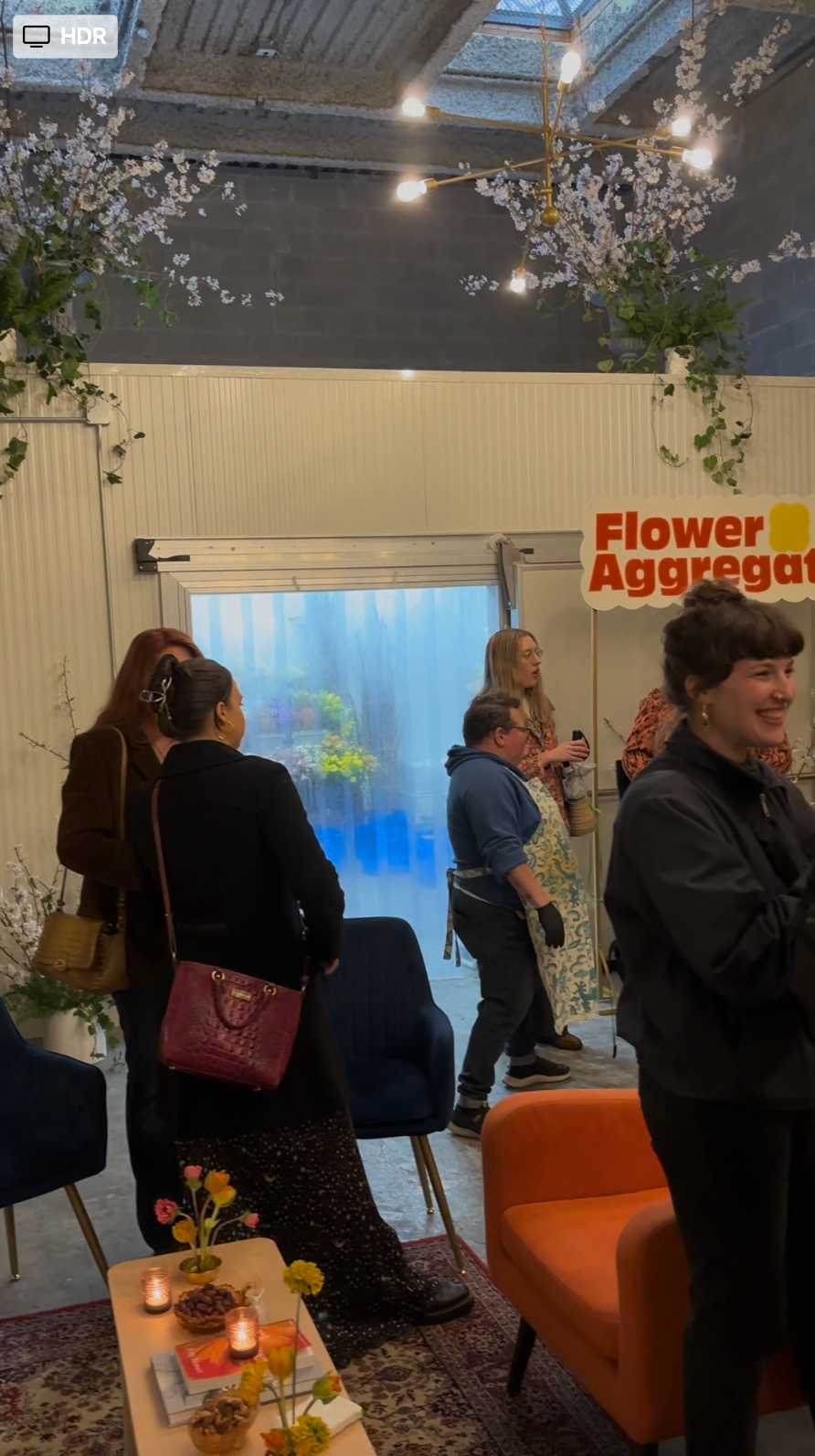 Group of people inside a flower shop, with a sign reading 'Flower Aggregator,' decorated with cherry blossom branches, ruffled curtains, and a coffee table with flowers, snacks, and candles.