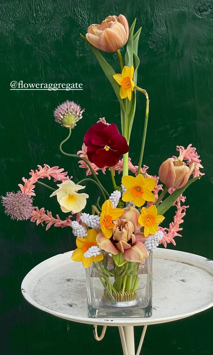 A glass vase with a colorful bouquet of various flowers, including tulips, daffodils, and other blooms, on a white round table against a dark green textured background.