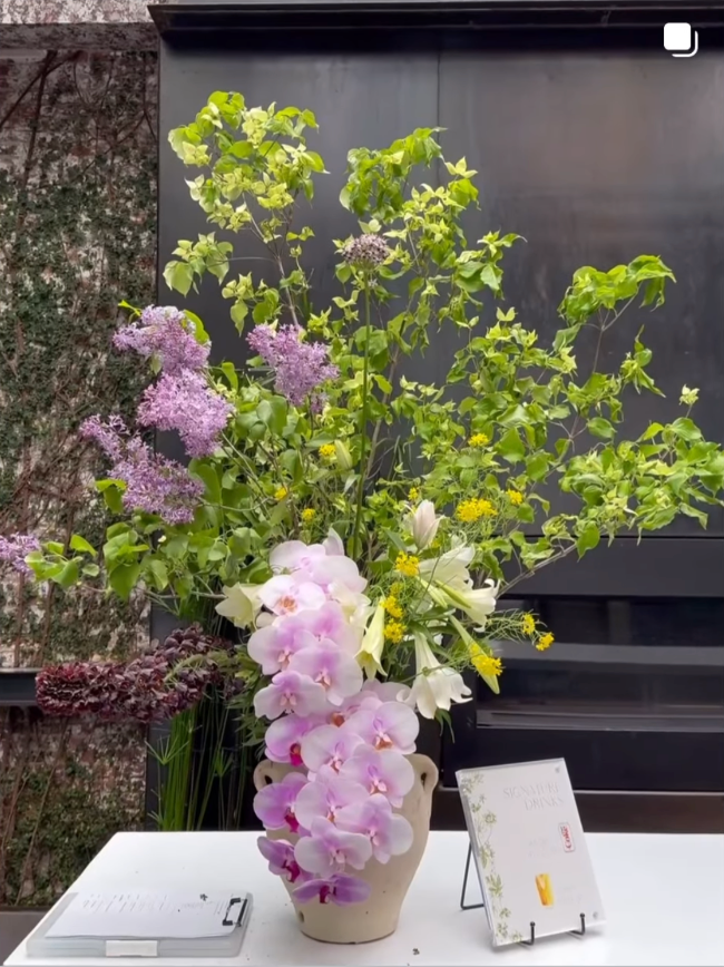 An elegant floral arrangement with pink orchids, white lilies, yellow flowers, and purple blossoms in a beige vase on a white table.