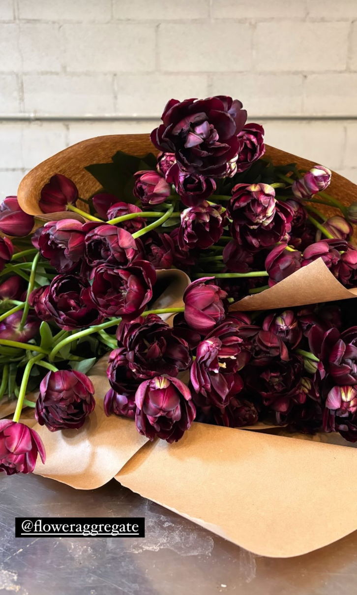 A bouquet of dark purple tulips wrapped in brown craft paper on a metallic surface, with a white brick wall background.