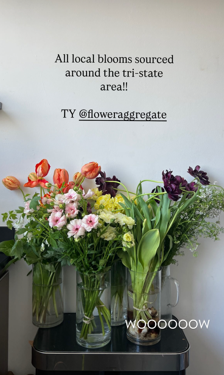 Four vases with a variety of colorful flowers, including pink, orange, purple, yellow, and white, are placed on a black table with a white wall behind. There is a sign on the wall that says, 'All local blooms sourced around the tri-state area!! TY @f
