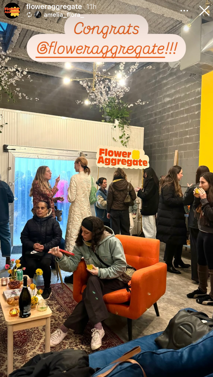 Indoor gathering with people socializing, some standing near a counter, others sitting on an orange chair. Decor includes flowers, lights, and a large sign that says 'Congrats @floweraggregate!!!'.
