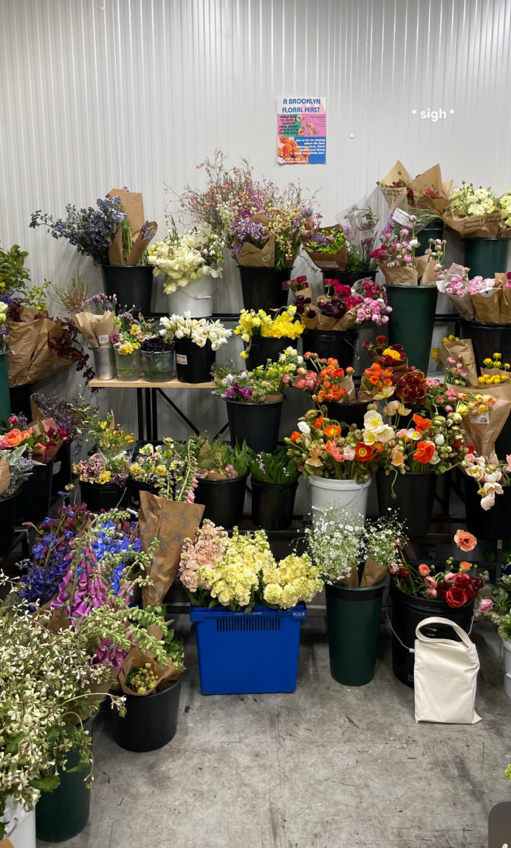 Multiple colorful flower bouquets and potted plants arranged on shelves and on the floor inside a floral shop. A pink flyer on the wall reads 'A Brooklyn Floral Feast.'