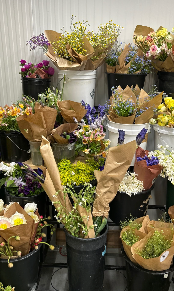 Various bouquets of fresh flowers wrapped in brown paper or in plastic containers, displayed on black and white buckets.