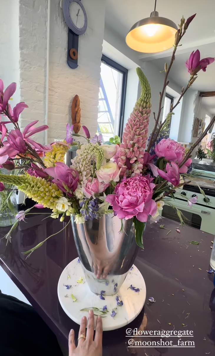 A large silver vase filled with pink, purple, white, and green flowers, including peonies, lupines, and magnolias, sits on a white round base on a dark table, with flower petals scattered around.