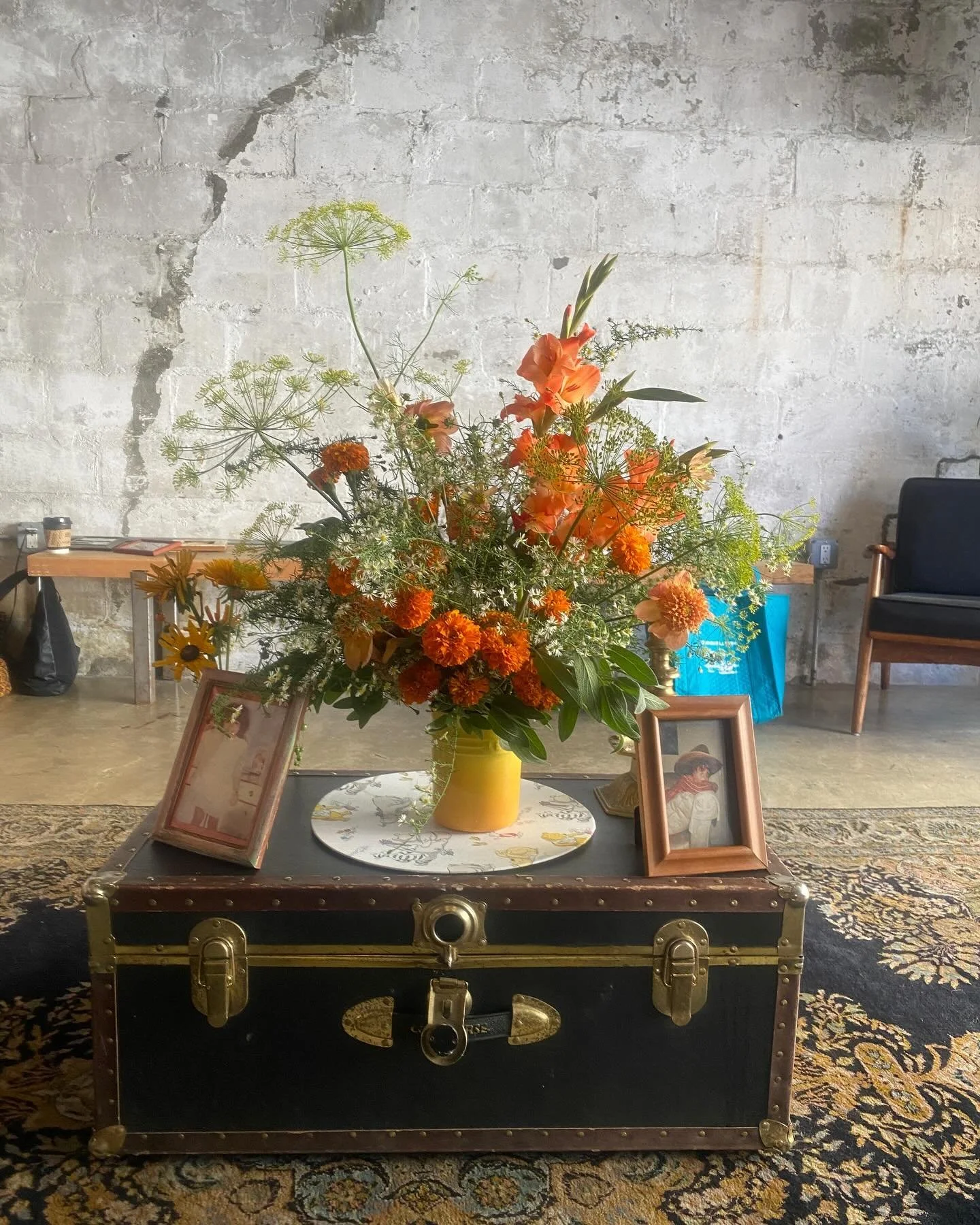 A large colorful flower arrangement on top of a vintage black trunk with gold hardware, placed on a patterned rug in front of a rustic concrete wall. Two framed photographs are also on the trunk.