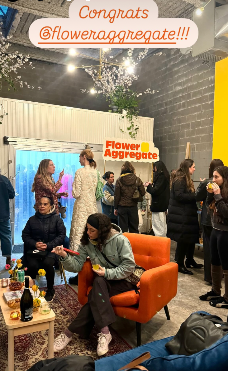 A group of people gathered indoors celebrating Flower Aggregate with a congratulatory message. Some are talking, others eating, with decorations and a sign that says "Flower Aggregate."