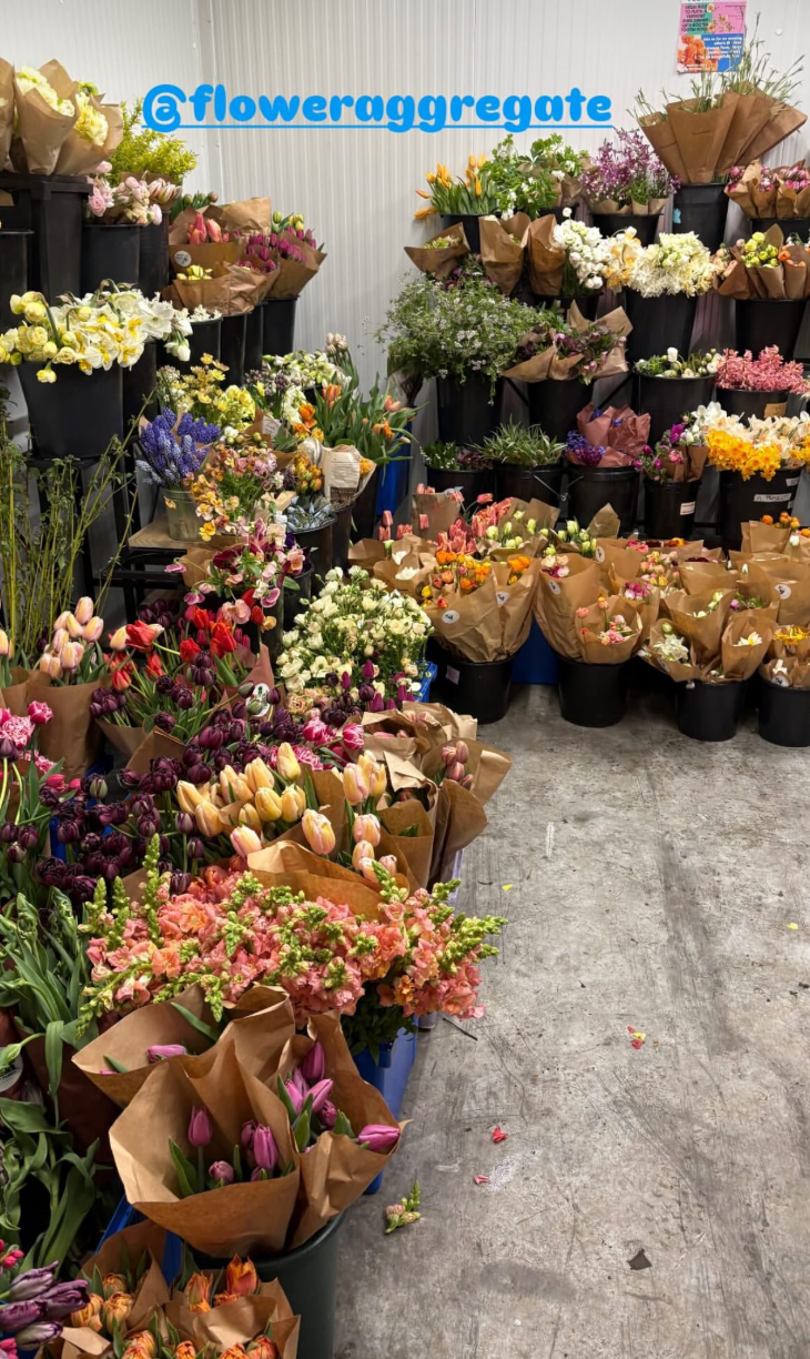A variety of flower bouquets in black pots and wrapped in brown paper bags at a flower shop, with a sign '@floweraggregate' in blue above them.