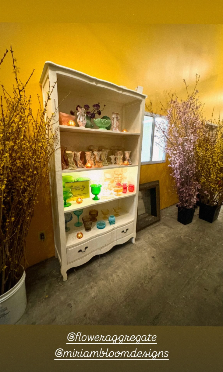 A white wooden display cabinet with three shelves filled with colorful glassware and decorative items, surrounded by tall branches with pink and yellow blossoms, set against a yellow wall inside a room.