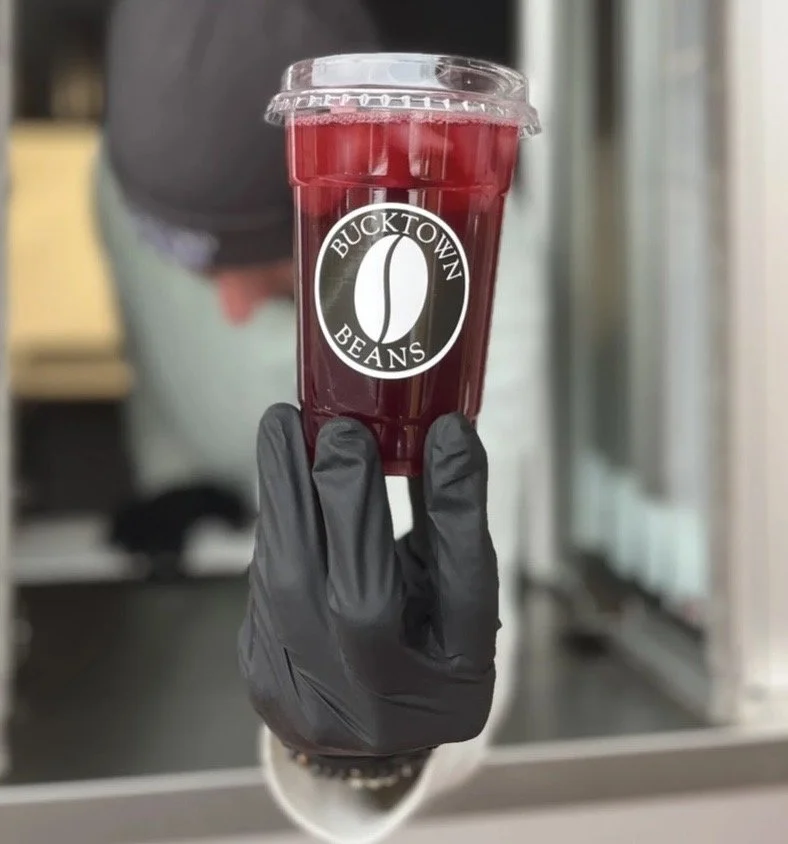 A hand wearing a black glove holding a clear plastic cup filled with a red beverage, with a Bucktown Beans logo on the side, and a clear plastic lid on top, in front of a blurred background.