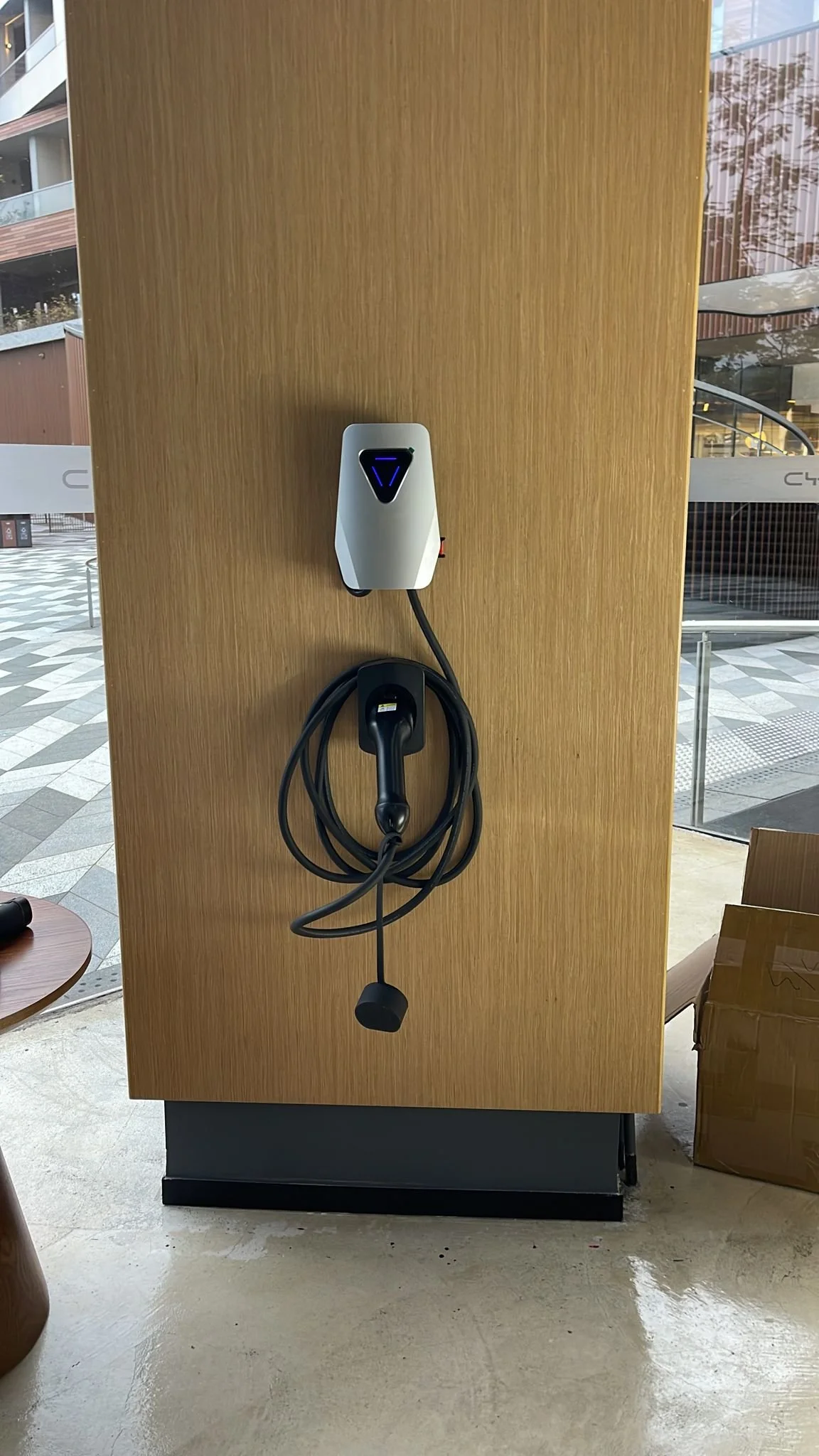 Electric car charging station mounted on a wooden panel indoors, with a cable and plug neatly stored below it.
