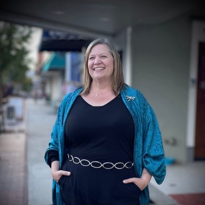 Smiling woman standing outdoors, wearing a black top, a blue cardigan, and a belt with circular links.