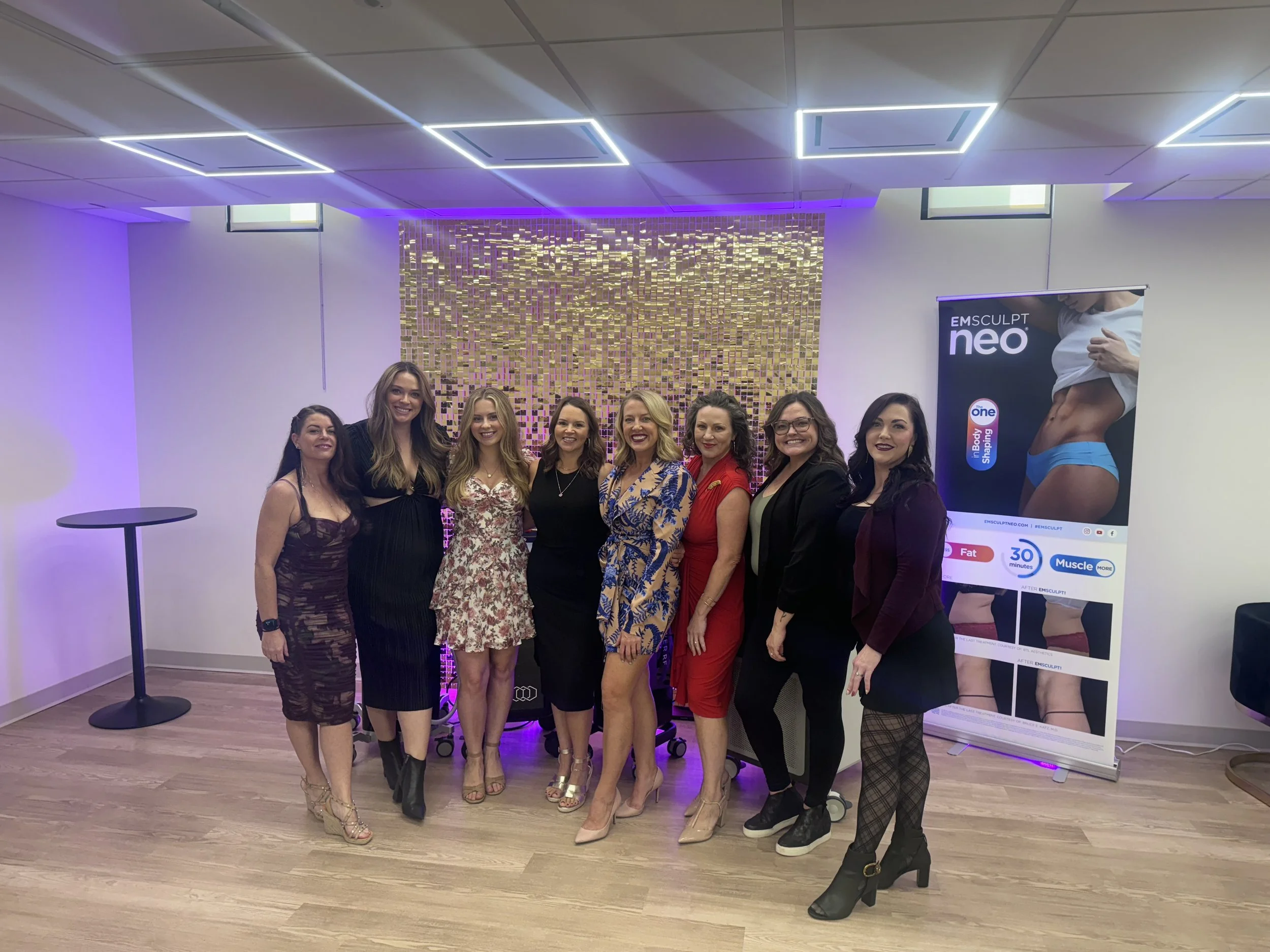 Group of women posing indoors in front of a banner for EMSCULPT neo, with a gold sequin backdrop and purple lighting. They are dressed in casual evening wear.