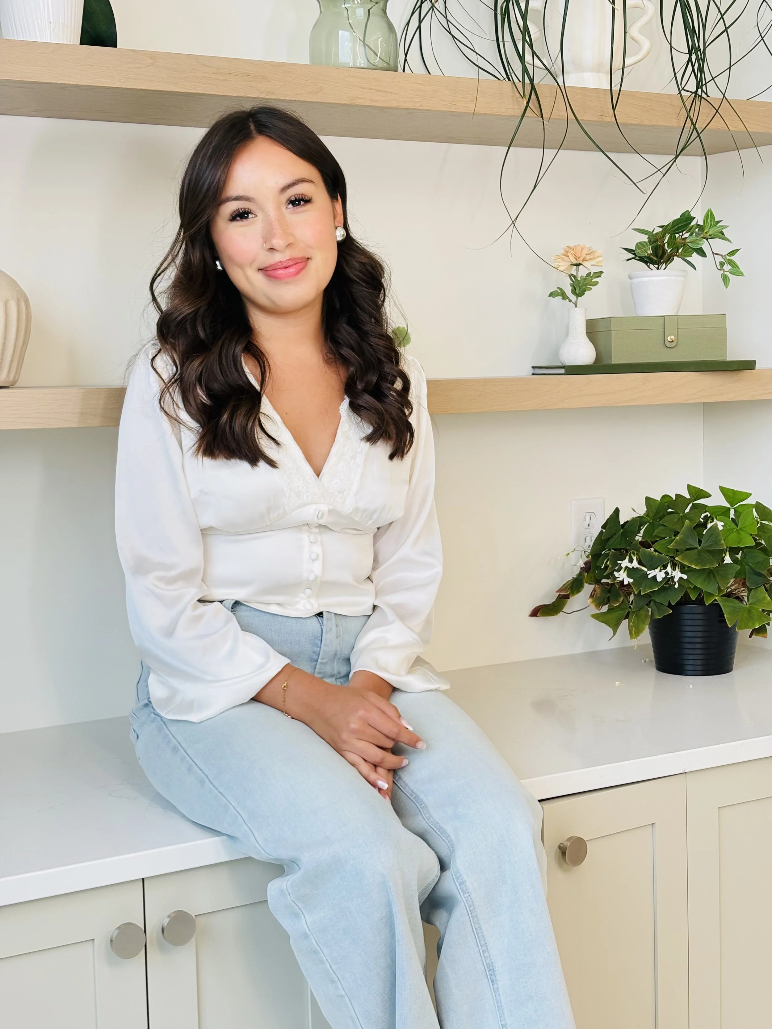 A young woman with long dark wavy hair, wearing a white blouse and light blue jeans, sitting on a white countertop in a room with light-colored walls and wooden shelves, surrounded by potted plants.