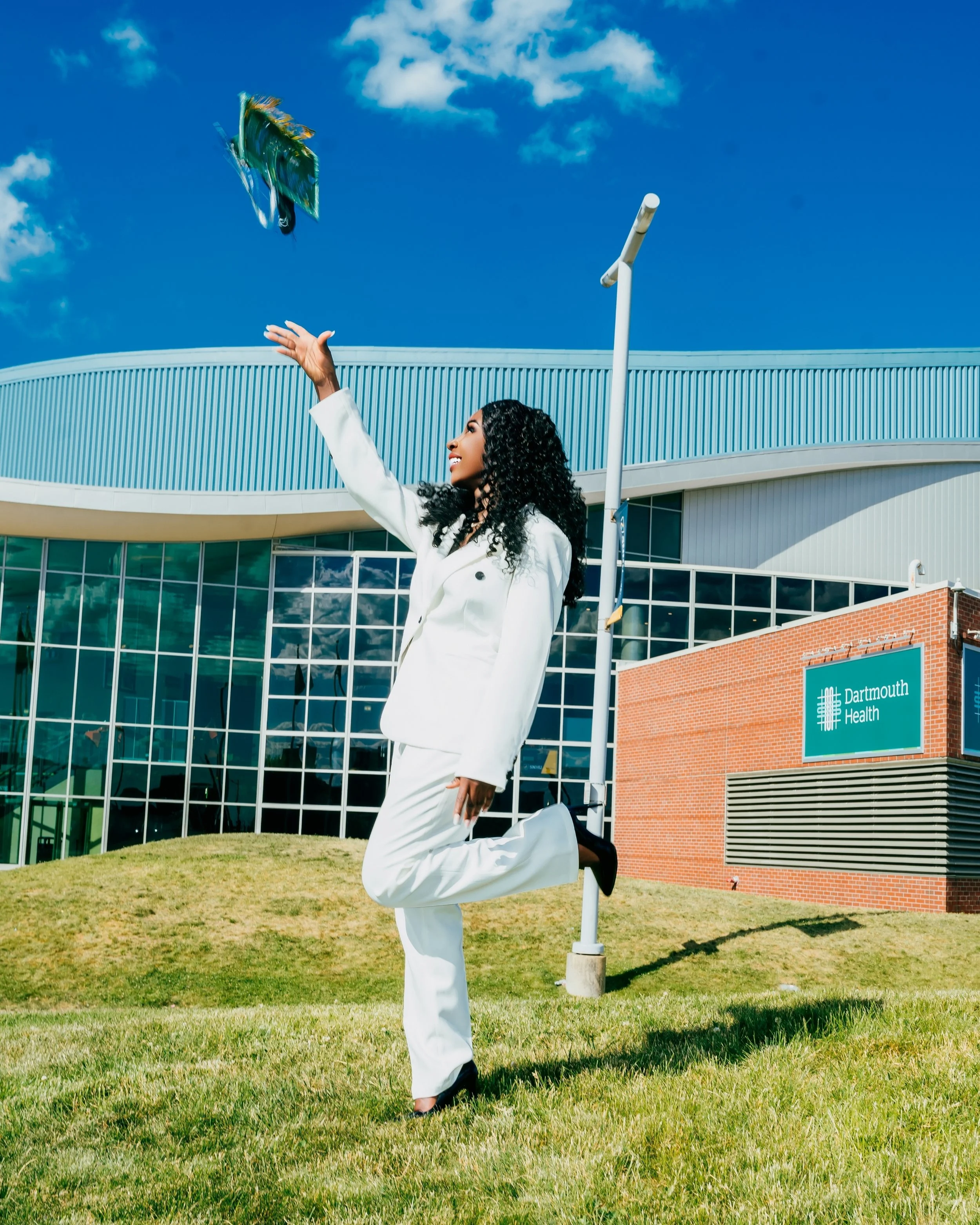 A woman in a white suit bouncing on one leg and catching a frisbee in front of Dartmouth Health building under a partly cloudy sky.