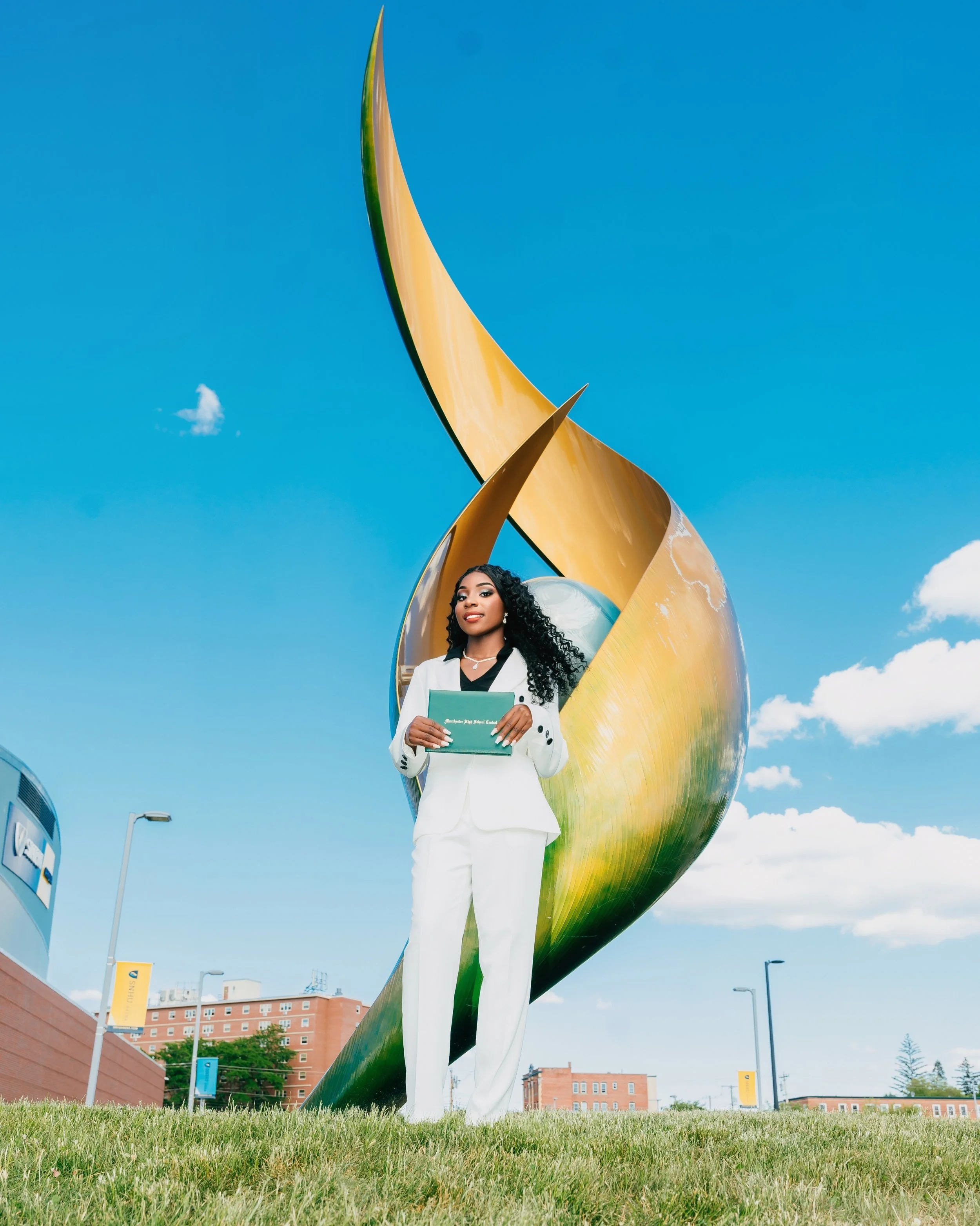 A woman with long curly black hair, dressed in a white suit holding a green folder, standing in front of an abstract outdoor sculpture with a clear blue sky overhead.
