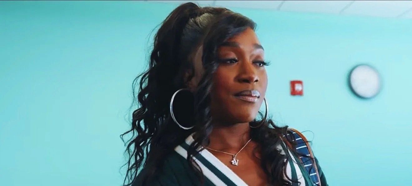 A woman with long curly black hair, wearing large hoop earrings, a necklace, and a striped shirt, standing in front of a turquoise wall with a clock.