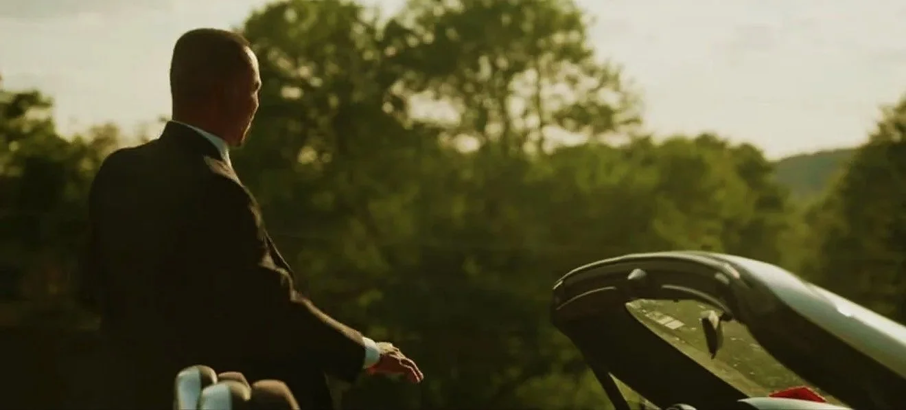 A man in a tuxedo standing next to a convertible car during sunset, with trees in the background.