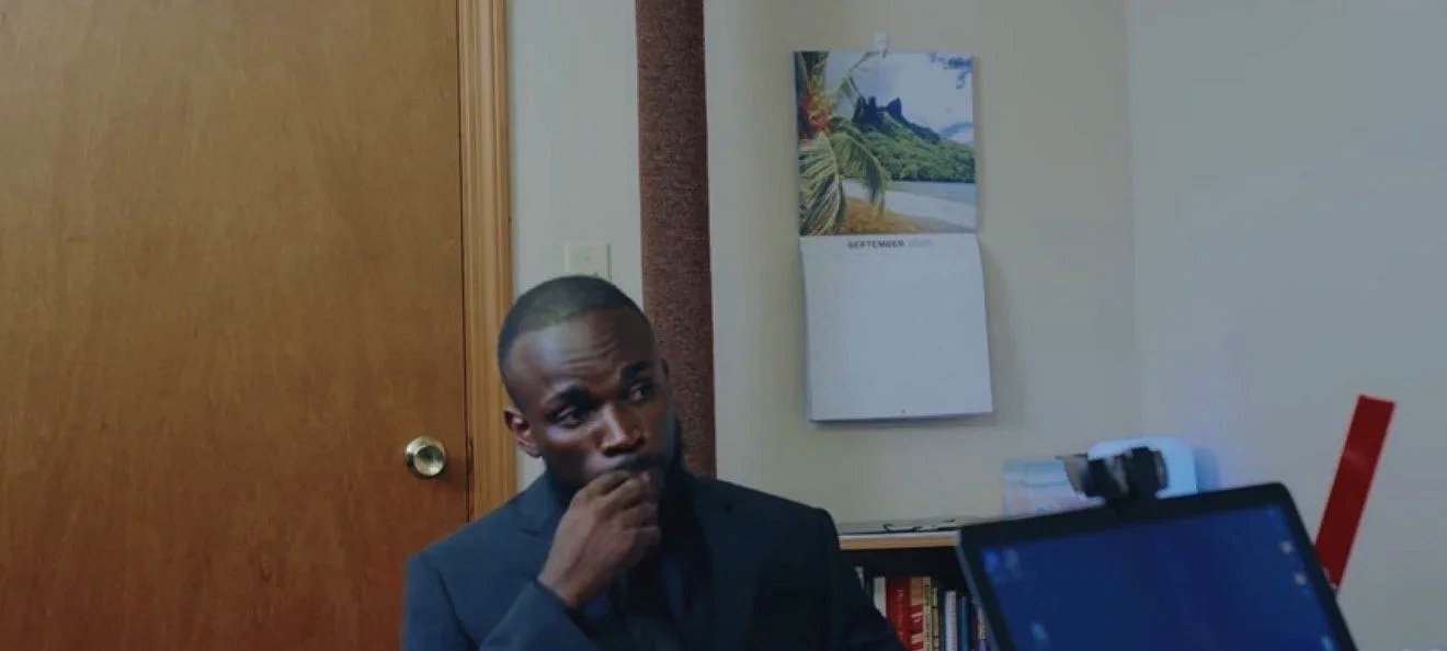 A man in a dark suit is sitting at a desk in an office room, with a calendar on the wall behind him and a stack of books to his right, while a laptop is in front of him and a webcam on top of it.