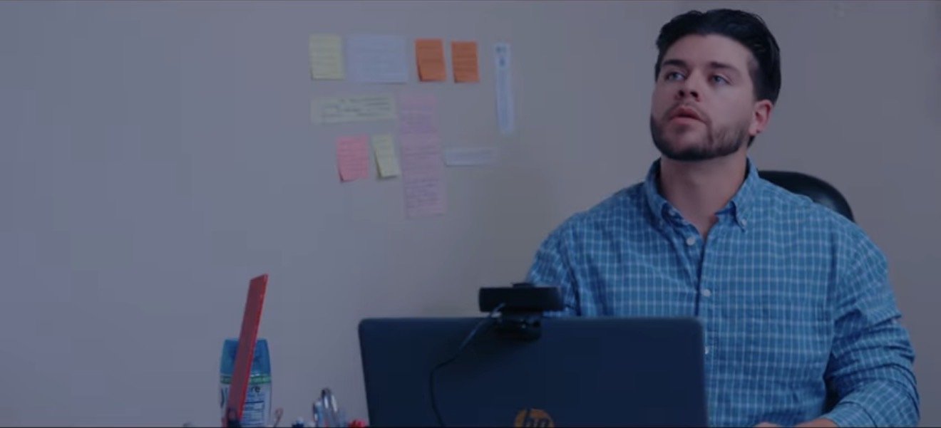 A man with a beard and glasses, wearing a blue checkered shirt, sitting at a desk with a laptop, in an office with sticky notes on the wall.