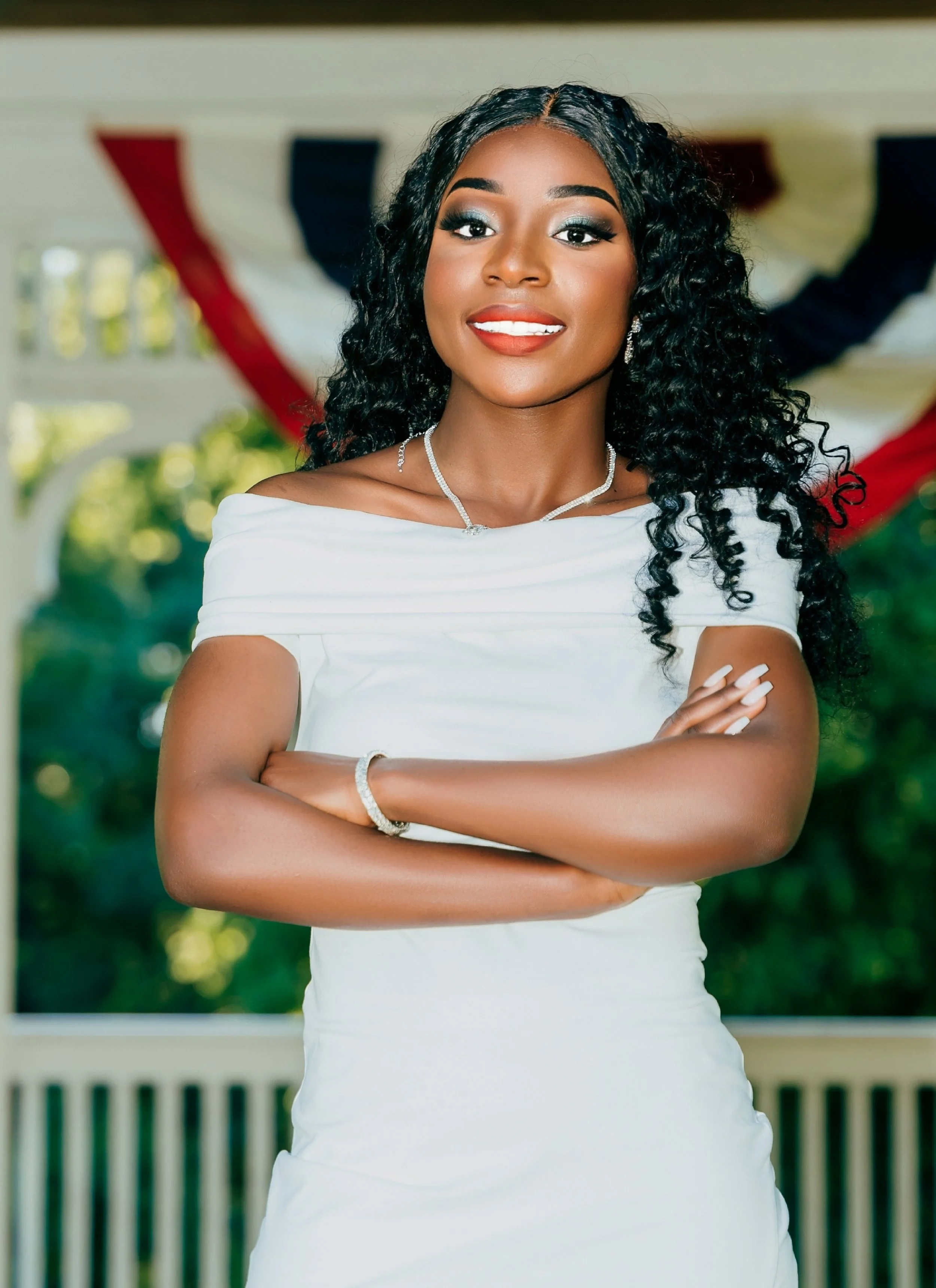 A young woman with dark curly hair, wearing a white dress and jewelry, smiling with arms crossed, with a blurred porch and greenery in the background.