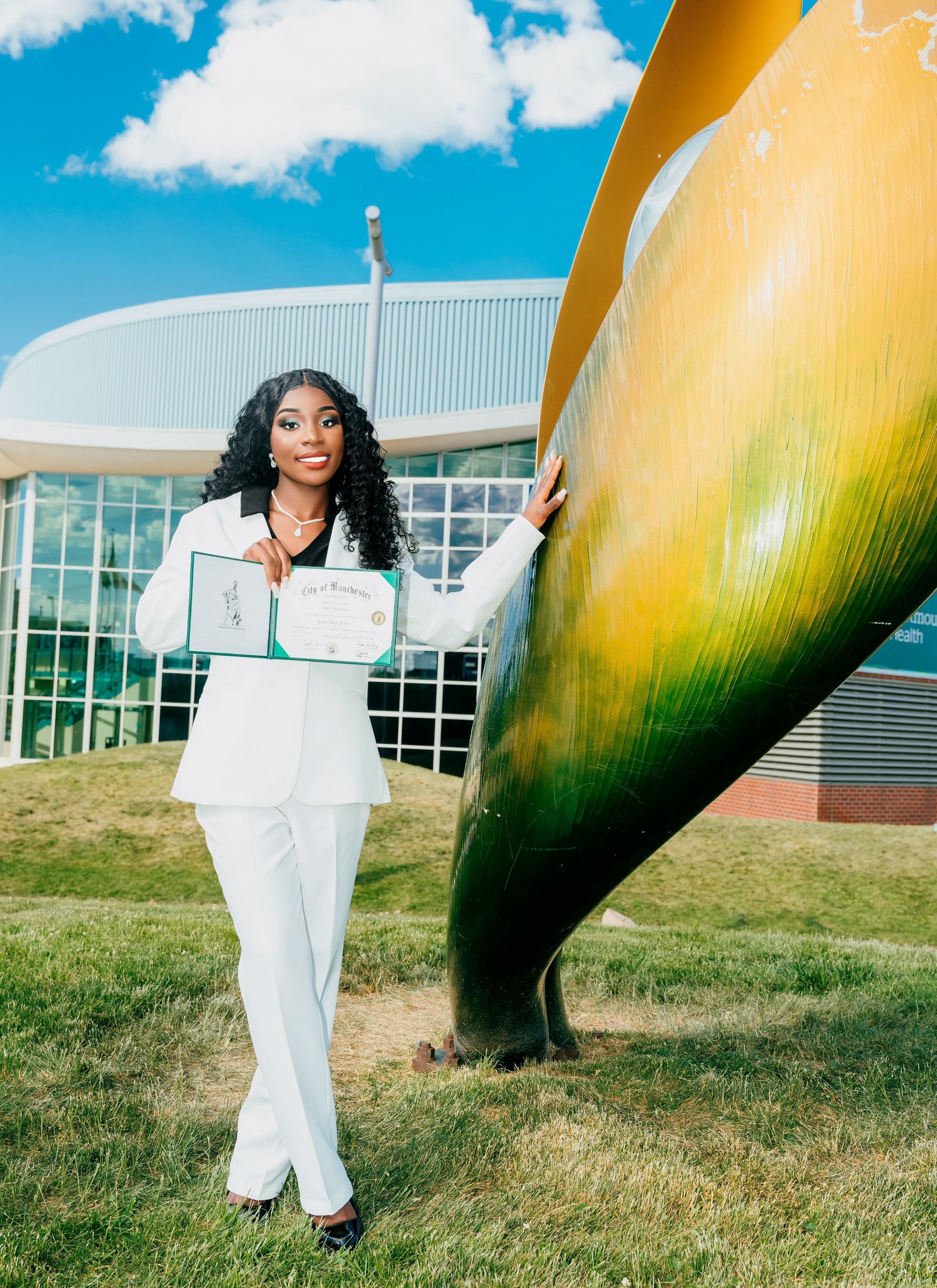 A woman in a white suit holding a certificate standing outdoors next to a large metallic sculpture with a modern building and blue sky in the background.