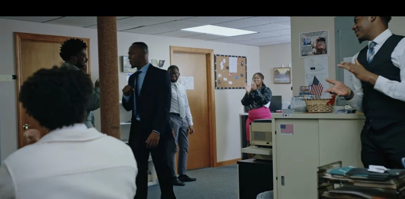 Office scene with five people, two men in suits are standing and talking, a woman in pink skirt is clapping, another woman in white is sitting facing away, and a man on the right is gesturing with hands.