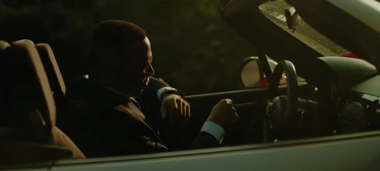 A man in a suit sitting in the driver's seat of a car at sunset, illuminated by the warm glow of the setting sun.