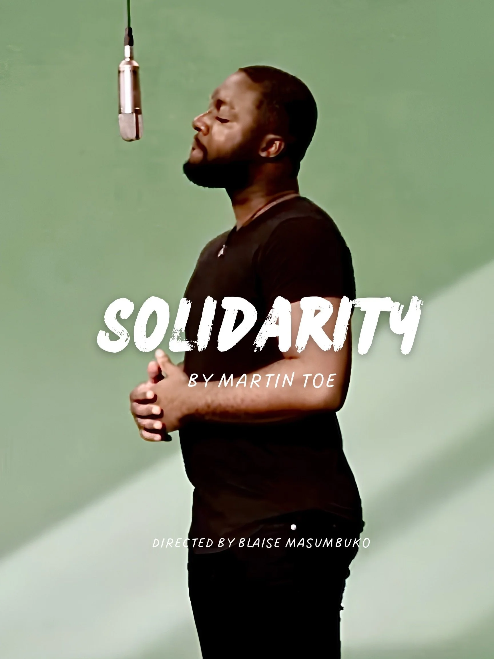 A man with a beard and short hair standing with clasped hands in front of his stomach in front of a green screen, with a microphone hanging above him. The image includes text indicating it is a solidarity video directed by Blaise Masumbuko.