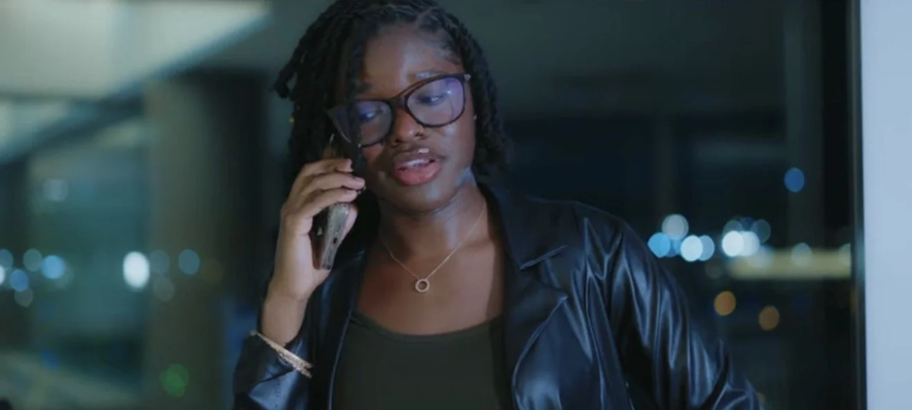 A woman with glasses and curly hair talking on a cell phone, wearing a black leather jacket, standing at night with city lights in the background.