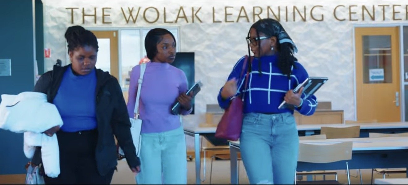 Three women walking and talking inside the Wolak Learning Center, a modern educational building with a reception area, tables, and chairs in the background.
