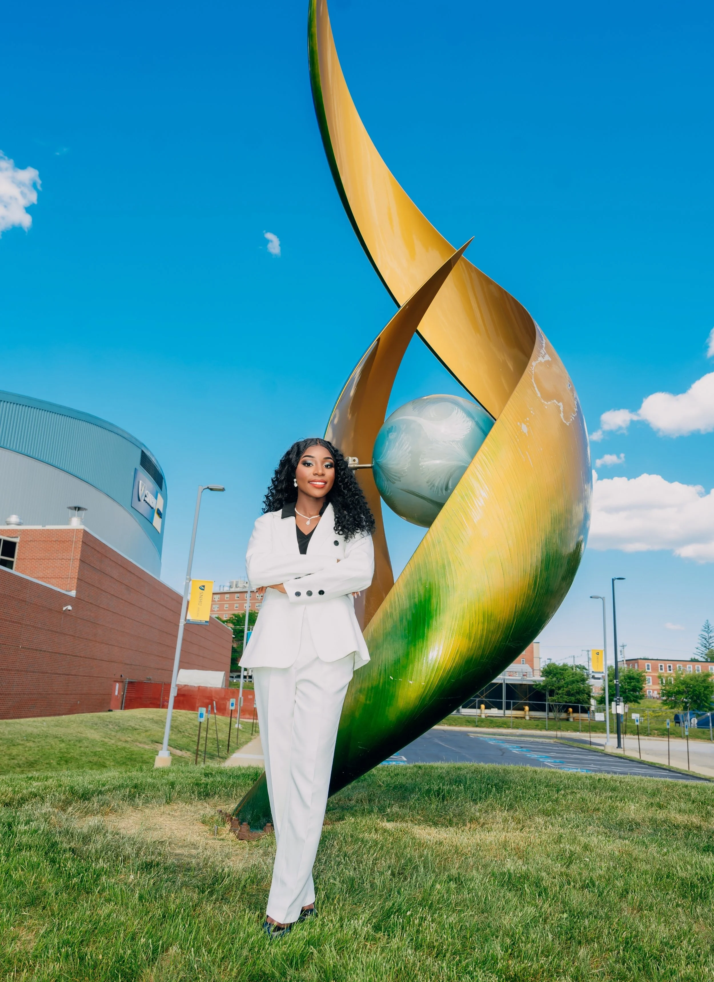 A woman in a white suit standing in front of a large golden and silver abstract sculpture outdoors, with a blue sky and some clouds in the background.