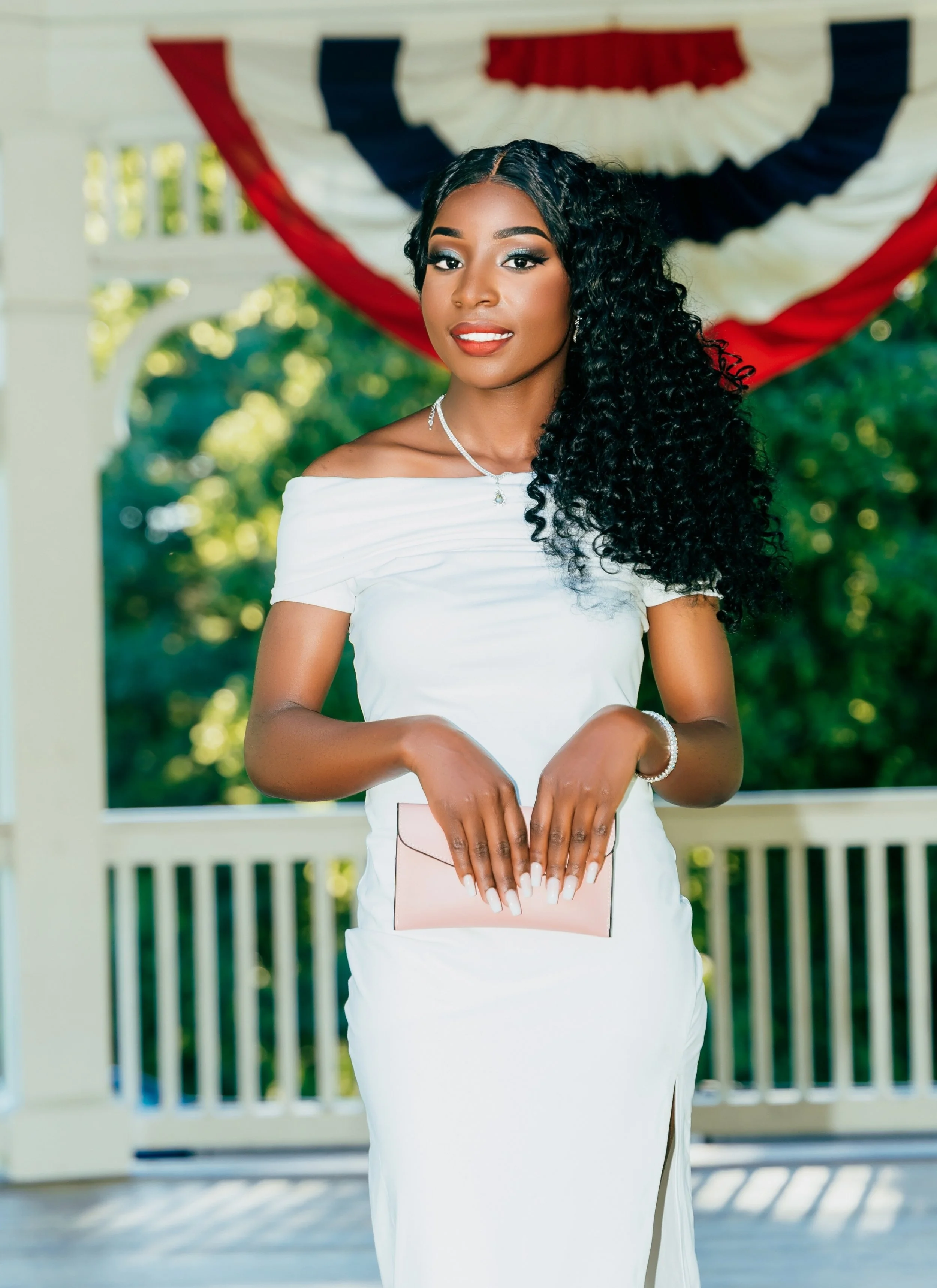 Young woman in a white dress holding a pink clutch purse, standing outdoors with a patriotic red, white, and blue bunting hanging above her.