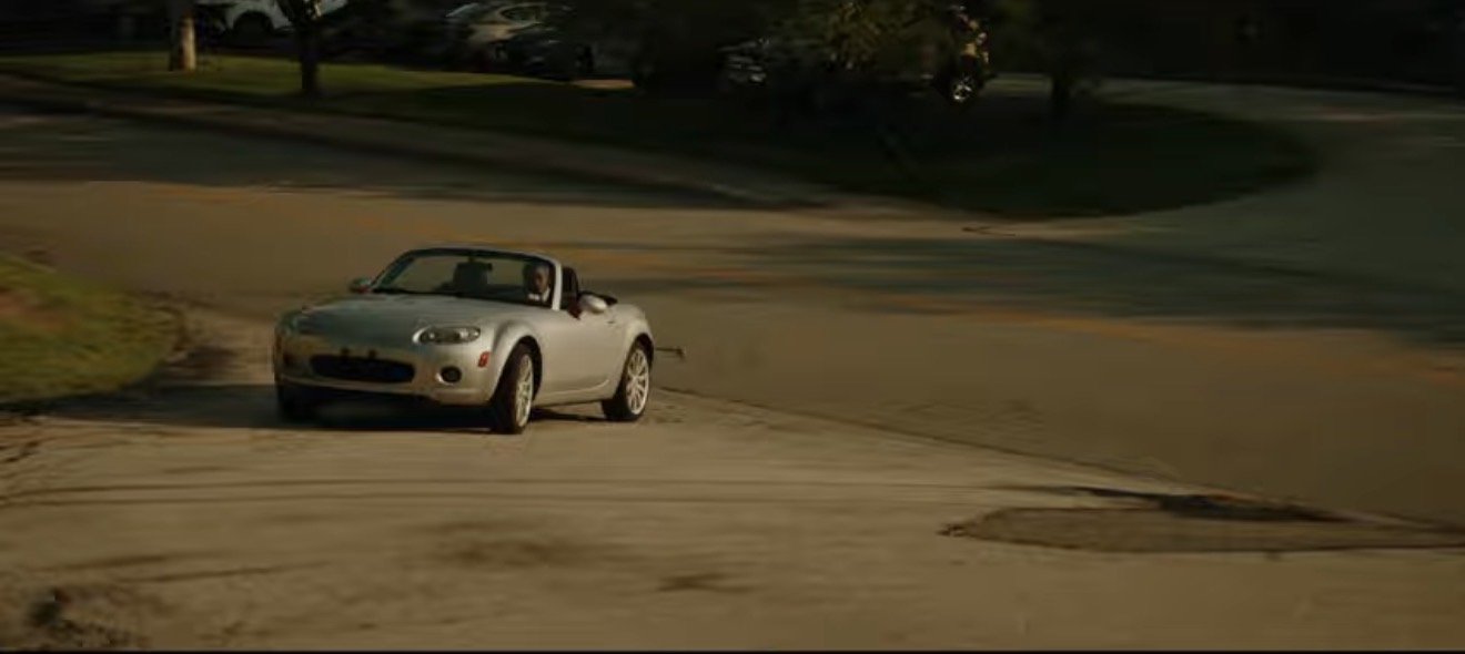 A silver convertible car driving on a residential street with trees and grass, with a person inside the car.