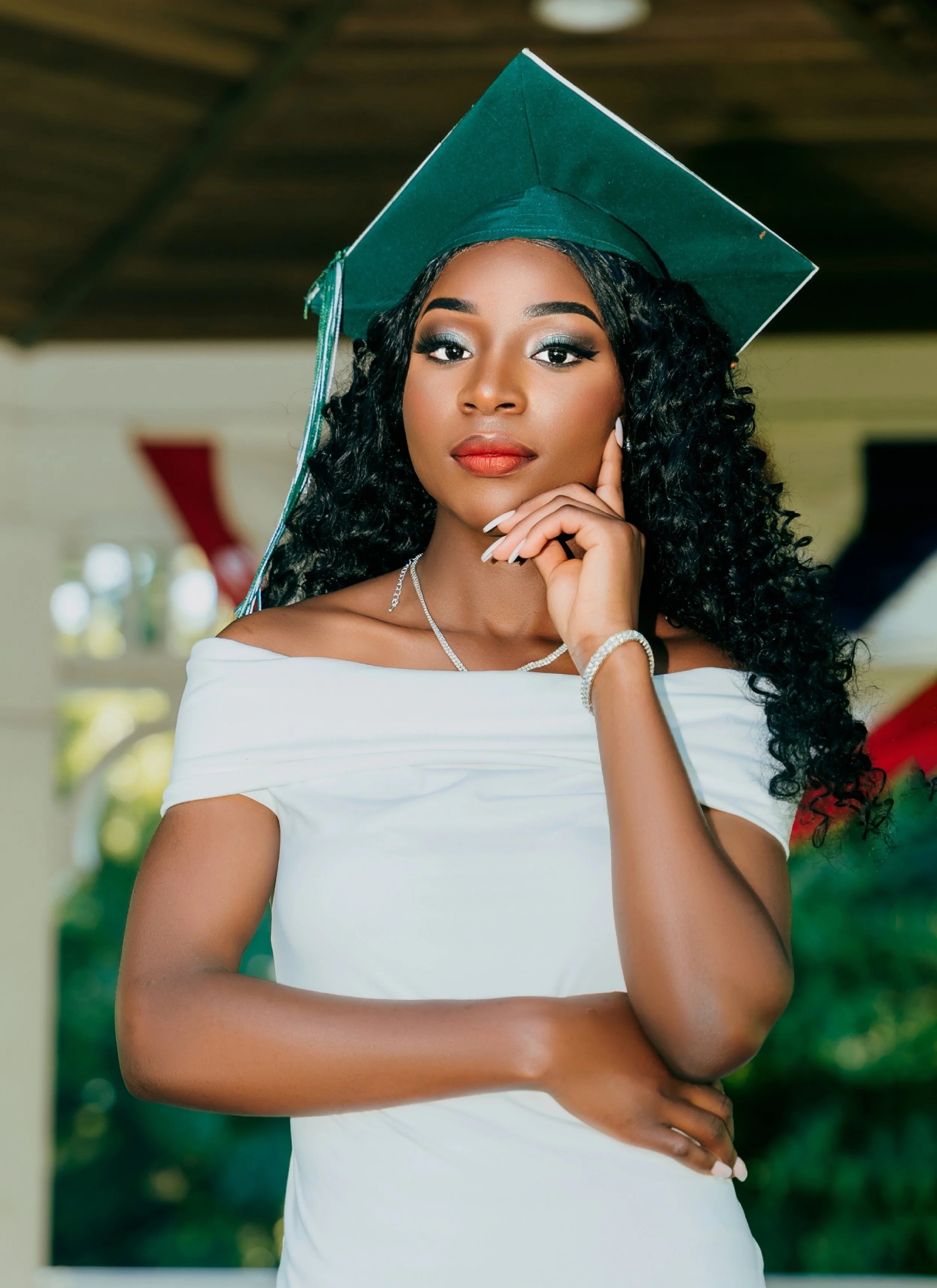 A young woman in a graduation cap and gown, standing outdoors with her hand resting on her chin, smiling gently.