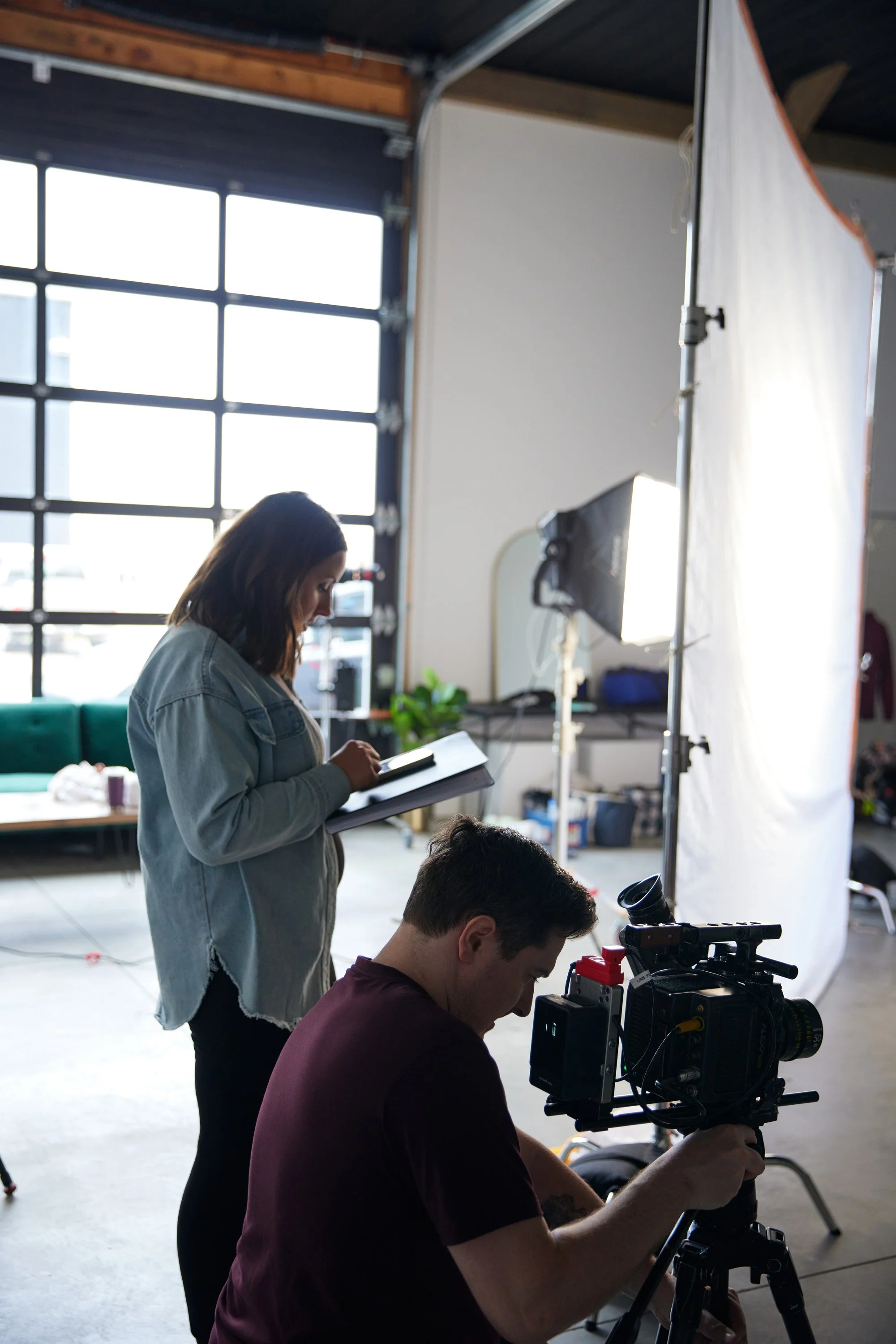 A woman and a man working on a film set; the woman is holding a clipboard and writing, while the man is operating a professional camera.