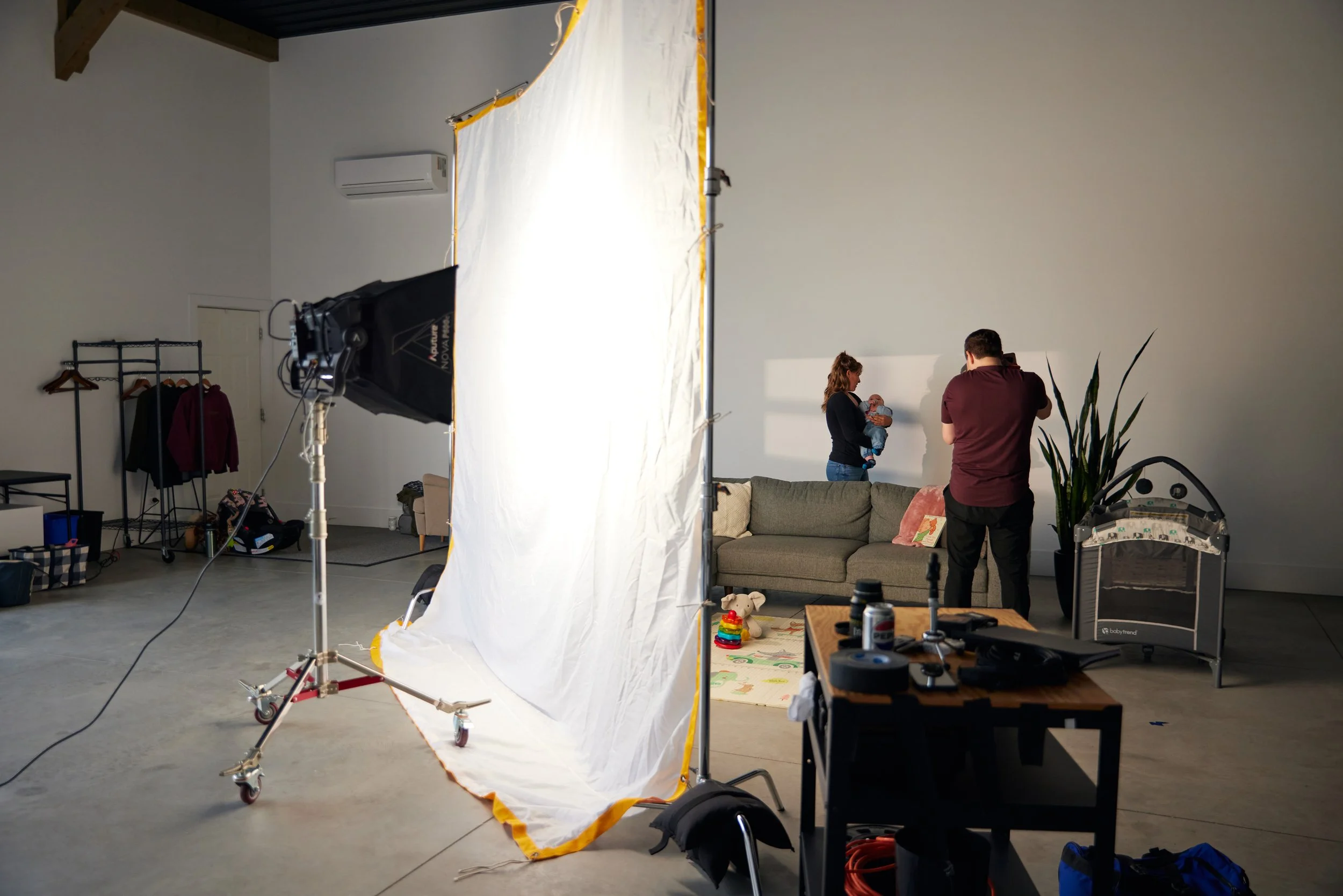 Indoor photoshoot setup with a white backdrop, a softbox lighting, a couch, a woman holding a baby, a photographer, and a baby crib in a spacious studio.