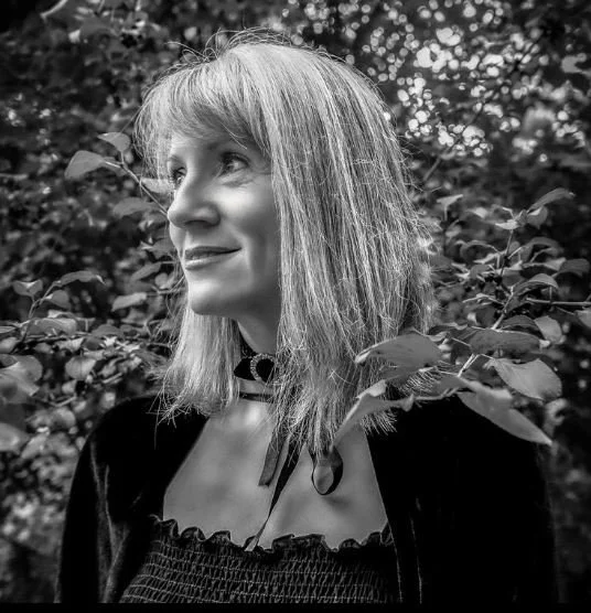 Black and white portrait of a woman with medium-length hair, wearing a black top and choker necklace, standing in front of leafy foliage.