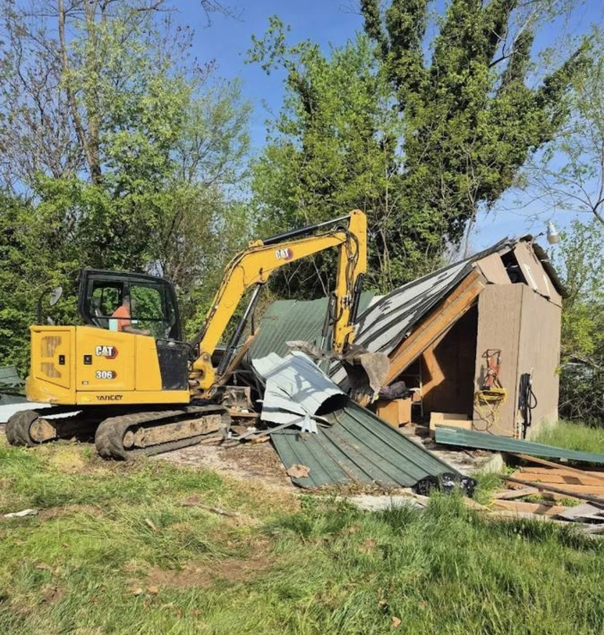 A yellow CAT mini excavator demolishing a small wooden shed with green metal roofing, surrounded by green grass and trees under a clear blue sky.
