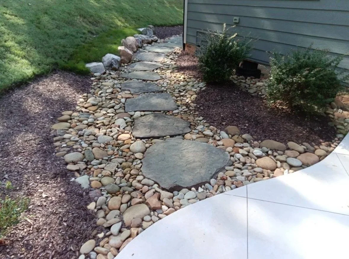 Stone pathway with large flat rocks and small pebbles, bordered by mulch and small bushes, next to a house with gray siding.