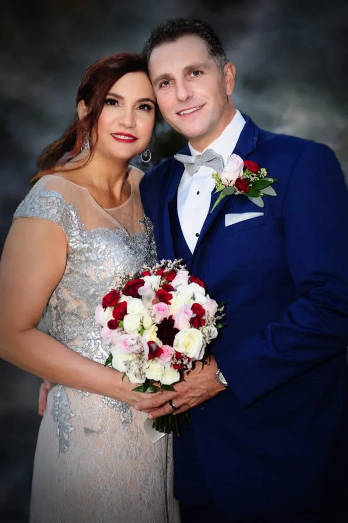 A couple posing for a wedding photo, with the man in a blue suit and bow tie, and the woman in a silver lace dress holding a bouquet of red, white, and pink flowers.