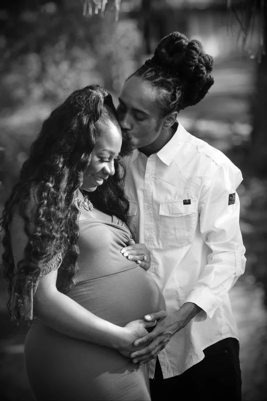 A man kissing a pregnant woman's forehead outdoors. The woman cradles her belly and smiles. It is a black and white photograph.