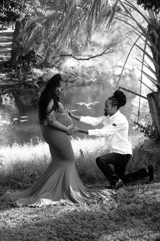 A black and white photo of a couple in a serene outdoor setting. The woman is pregnant and wearing a long dress, while the man is kneeling in front of her, holding her hands.