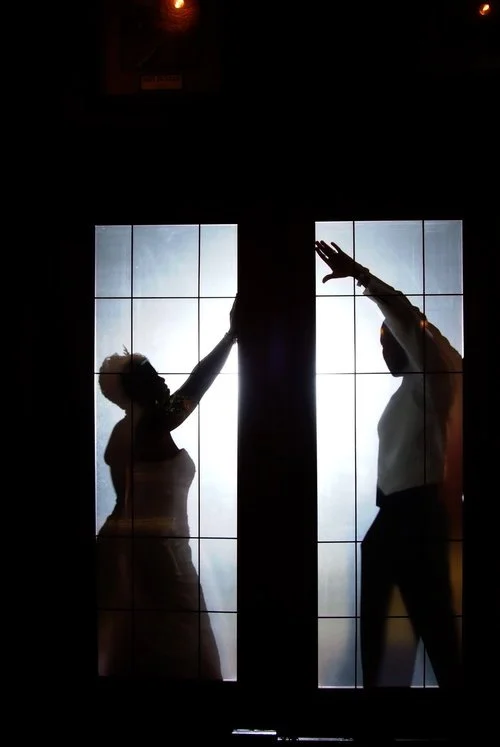 Silhouettes of a couple in wedding attire behind frosted glass doors, reaching towards each other.