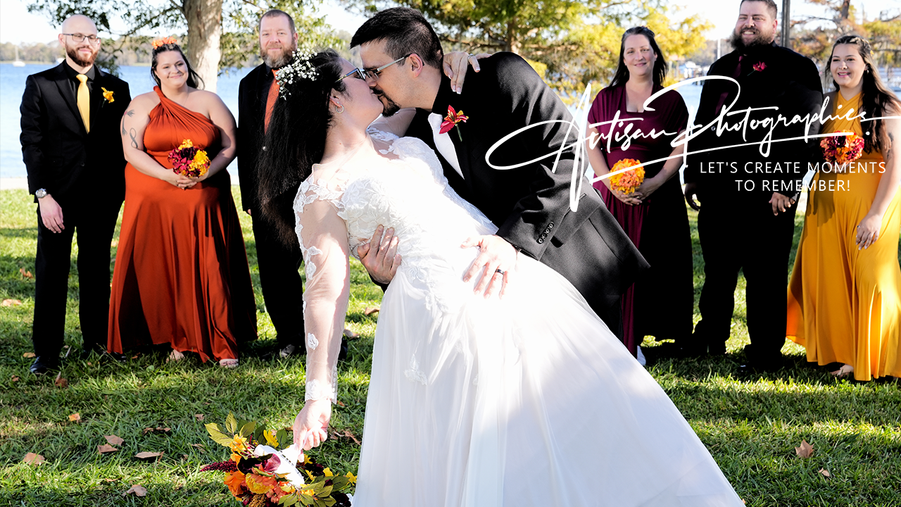 A wedding couple kissing outdoors with a bridal party in the background. The bride is in a white gown, and the groom is in a black suit. The bridal party wears coordinated dresses and suits. The scene is relaxed and joyful, set on a grassy area near a body of water.