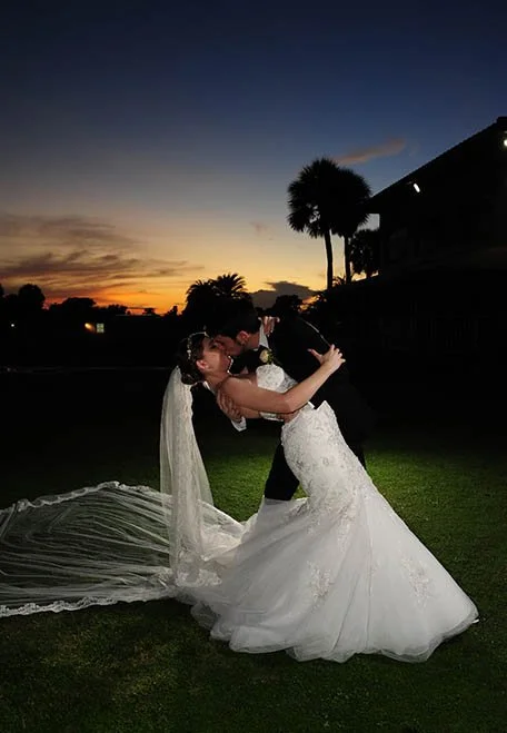 Bride and groom in wedding attire sharing a romantic dance at sunset on a grassy lawn.