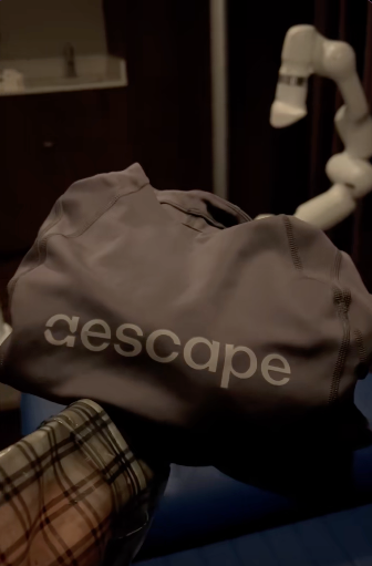 A gray bag with the word 'escape' printed on it, held by a person in a plaid shirt, in a room with a robotic arm in the background.