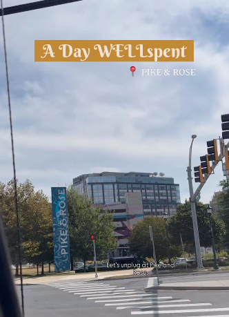 City street scene with a pedestrian crossing, traffic lights, trees, and a modern building. Overlaid text reads 'A Day WELLspent' and 'PIKE & ROSE' with a small map pin icon, and a caption at the bottom says 'Let's unplug at Pike & Rose'.