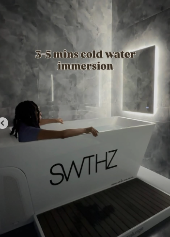 A woman relaxing in a white bathtub labeled 'SWTHZ' in a modern bathroom with gray marble walls and a window.