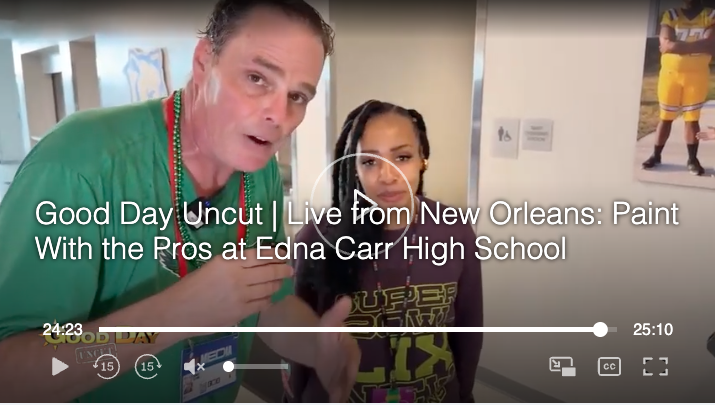 A man in a green shirt and a woman with braided hair stand indoors, with framed artwork and a sports poster on the wall behind them, during a video recording.